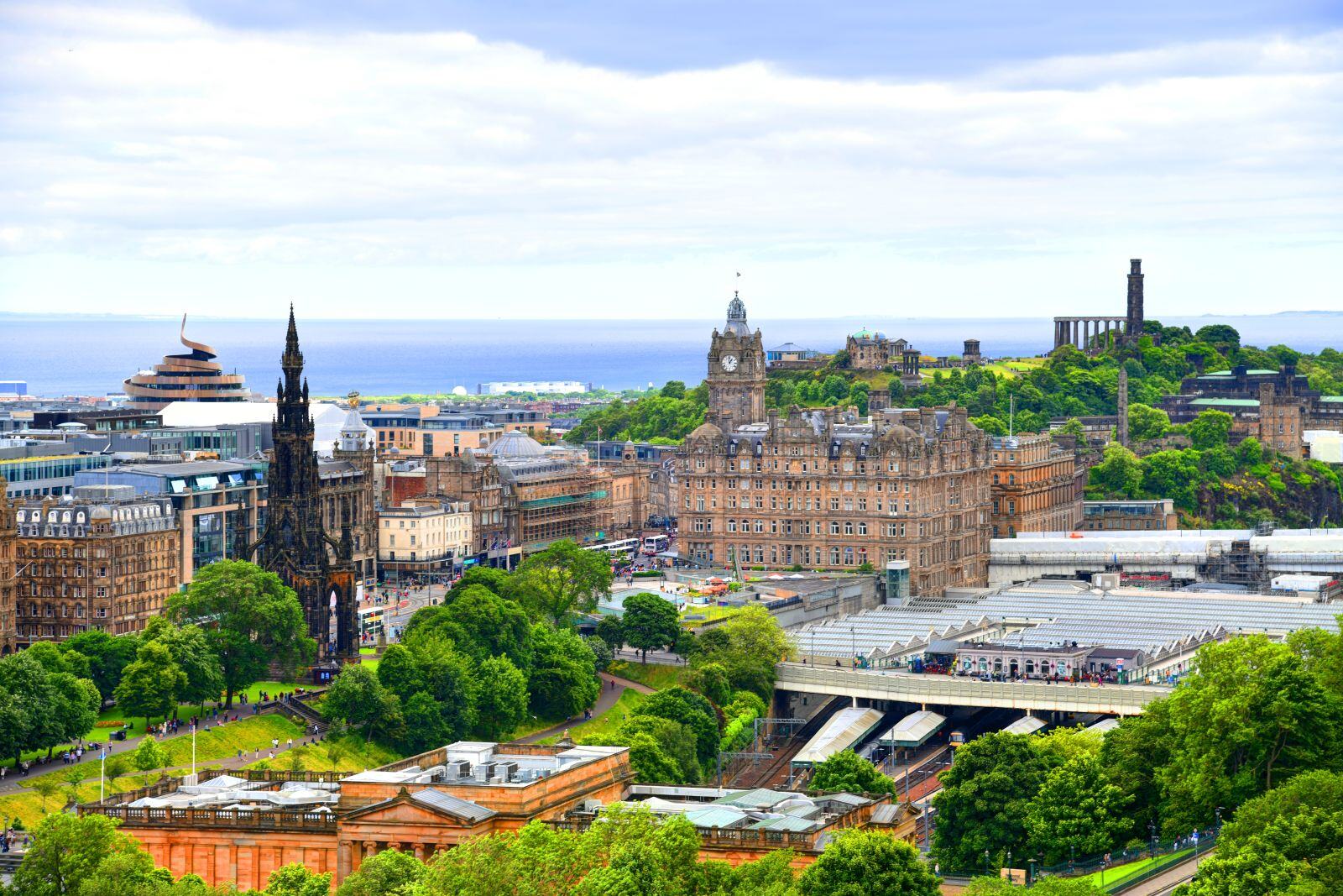 Panoramic view of Edinburgh, Scotland, showcasing a blend of historic and modern architecture. The Scott Monument, a tall Gothic spire, stands on the left, while the Balmoral Hotel with its clock tower is centrally located. Calton Hill rises in the background with several monuments. The foreground features lush green trees and gardens, and the middle ground is filled with buildings and busy streets. The North Sea is visible in the distance beneath a partly cloudy sky.