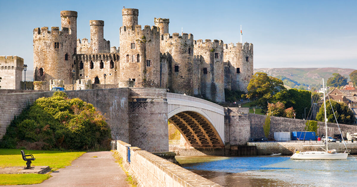 Historic castle with multiple towers and battlements beside a body of water, connected to land by a stone bridge. A sailboat is docked nearby, and the area is surrounded by greenery, trees, and distant buildings. An empty bench sits along a pathway leading toward the castle.