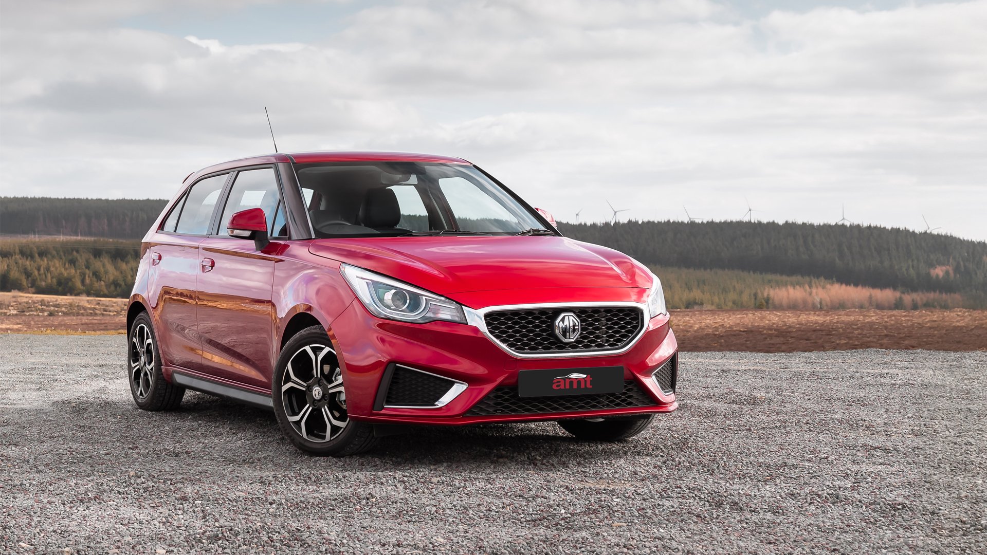 Red MG3 hatchback parked on a rural gravel road with scenic forest and hills in the background.