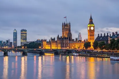 View of the Palace of Westminster and Big Ben in London at dusk, seen from across the River Thames. The buildings are warmly lit and reflected in the calm water. The Elizabeth Tower stands prominently on the right, while Westminster Bridge stretches across the river to the left. Modern buildings and a cloudy sky form the background.