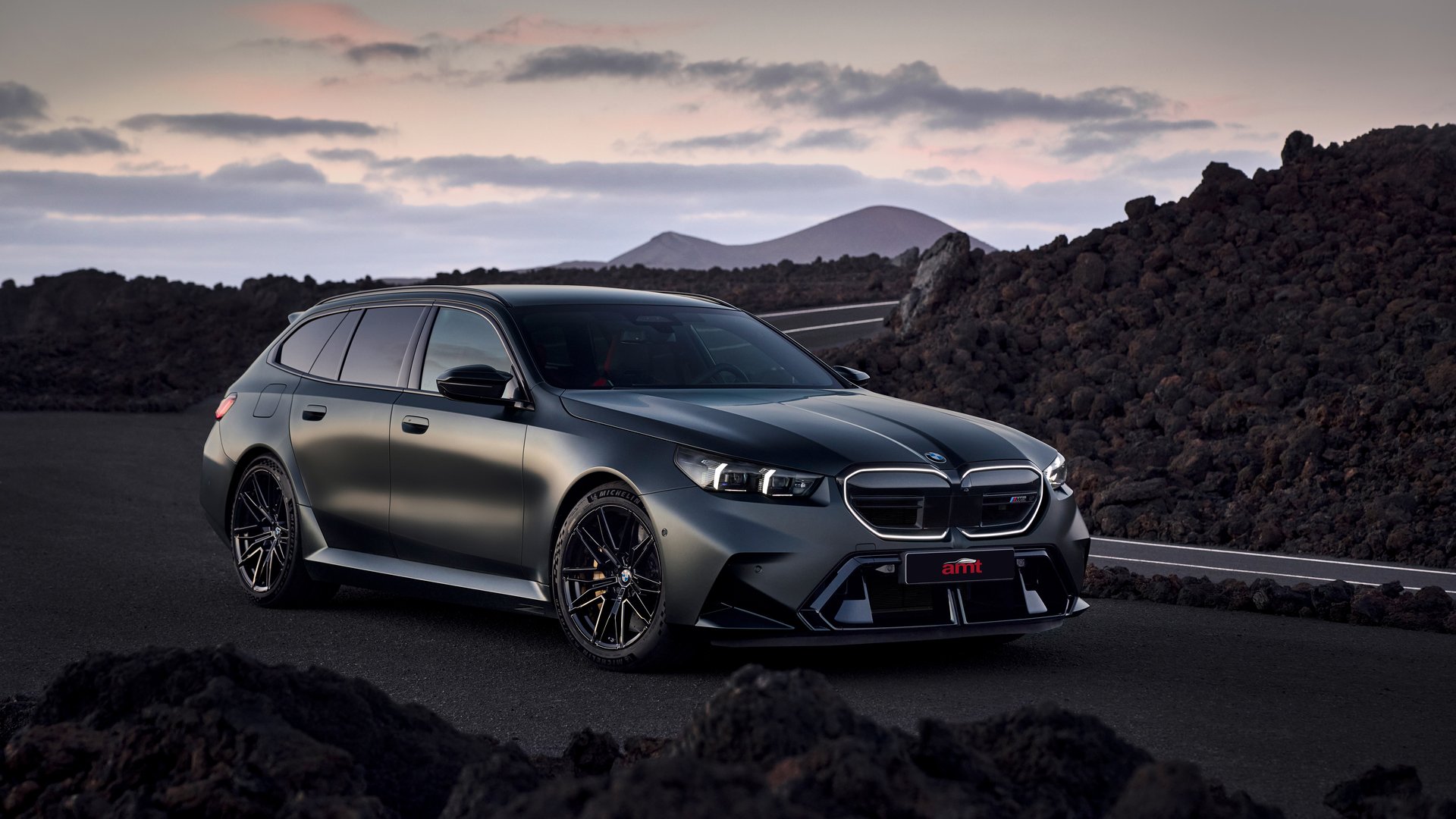 BMW M5 Touring estate in matte black driving through mountain landscape at sunset.